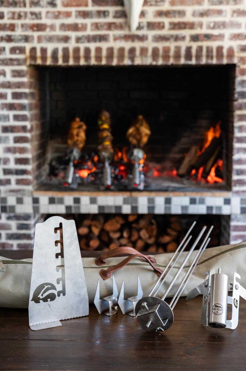 Chicken pieces turning on skewers in a brick fireplace with stacked firewood behind