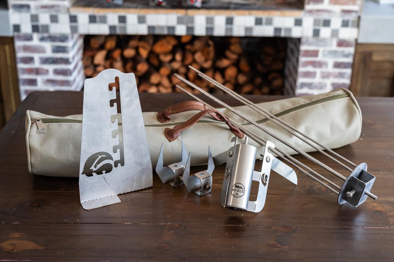 The full SpitAlles kit laid out on a table in front of a wood store: canvas bag, counter stand, skewers and motor